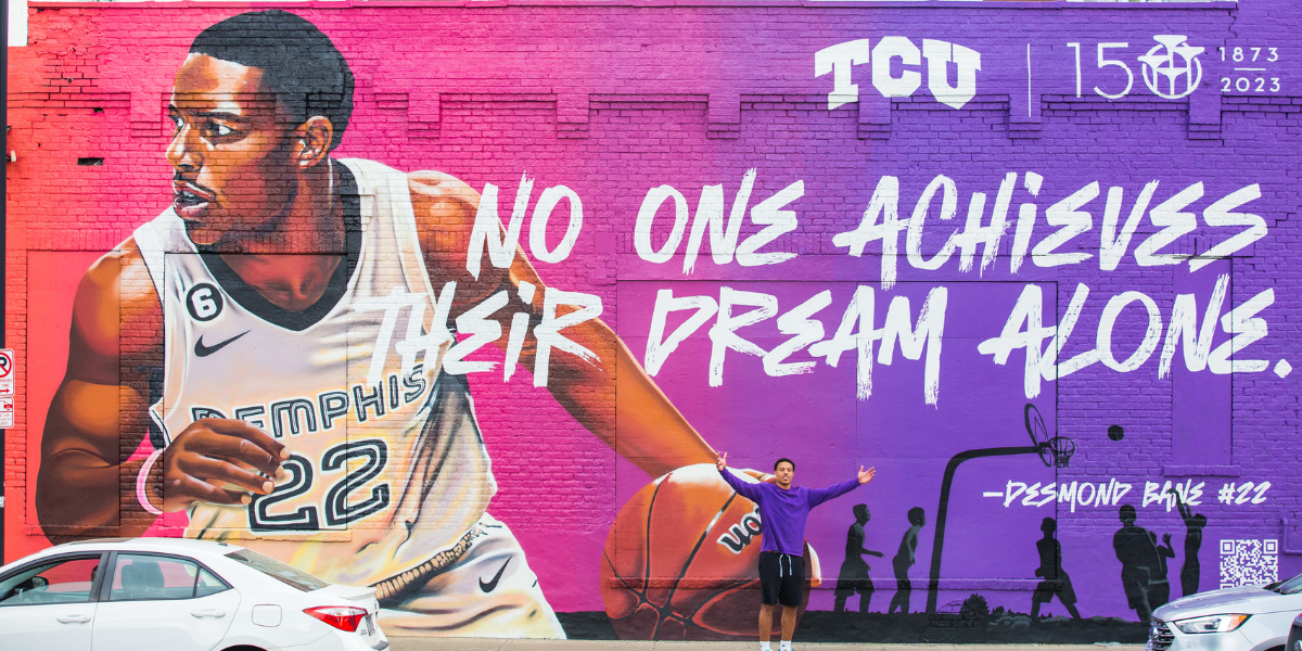 Desmond Bane standing with his arms spread in front of a mural. The mural has a pink to purple ombre background and shows Bane in his Memphis basketball unfirom holding a basketball. In the top left corner are the TCU logo and the TCU 150th anniversary logo. In the middle of the mural is the quote "No one achieves their dream alone" attributed to Desmond Bane.