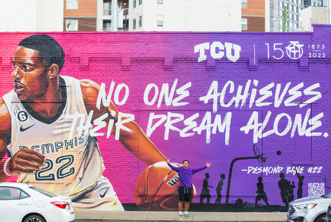 Desmond Bane standing with his arms spread in front of a mural. The mural has a pink to purple ombre background and shows Bane in his Memphis basketball unfirom holding a basketball. In the top left corner are the TCU logo and the TCU 150th anniversary logo. In the middle of the mural is the quote 