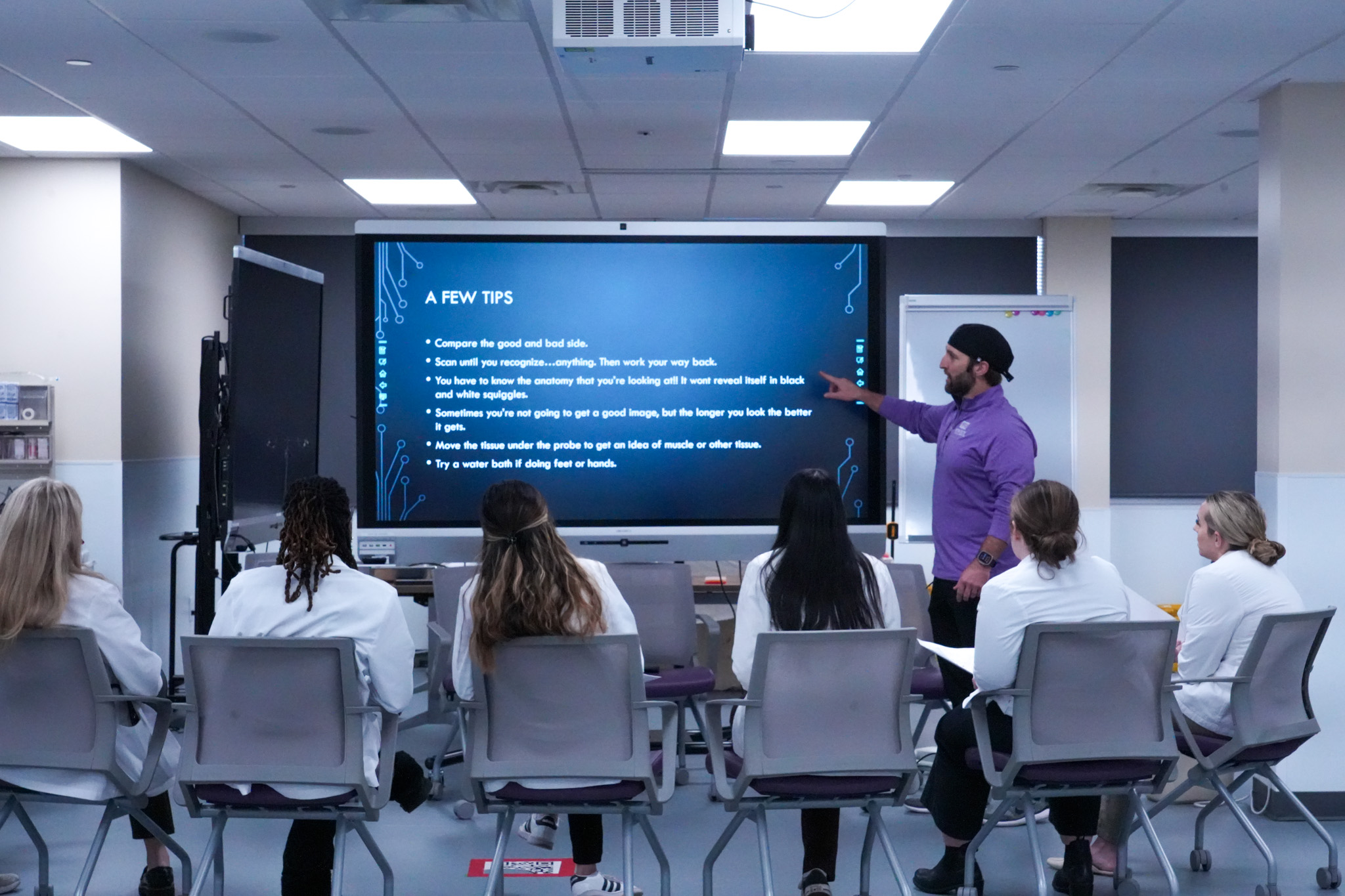 instructor teaching while pointing at projector screen in front of students in white coats.