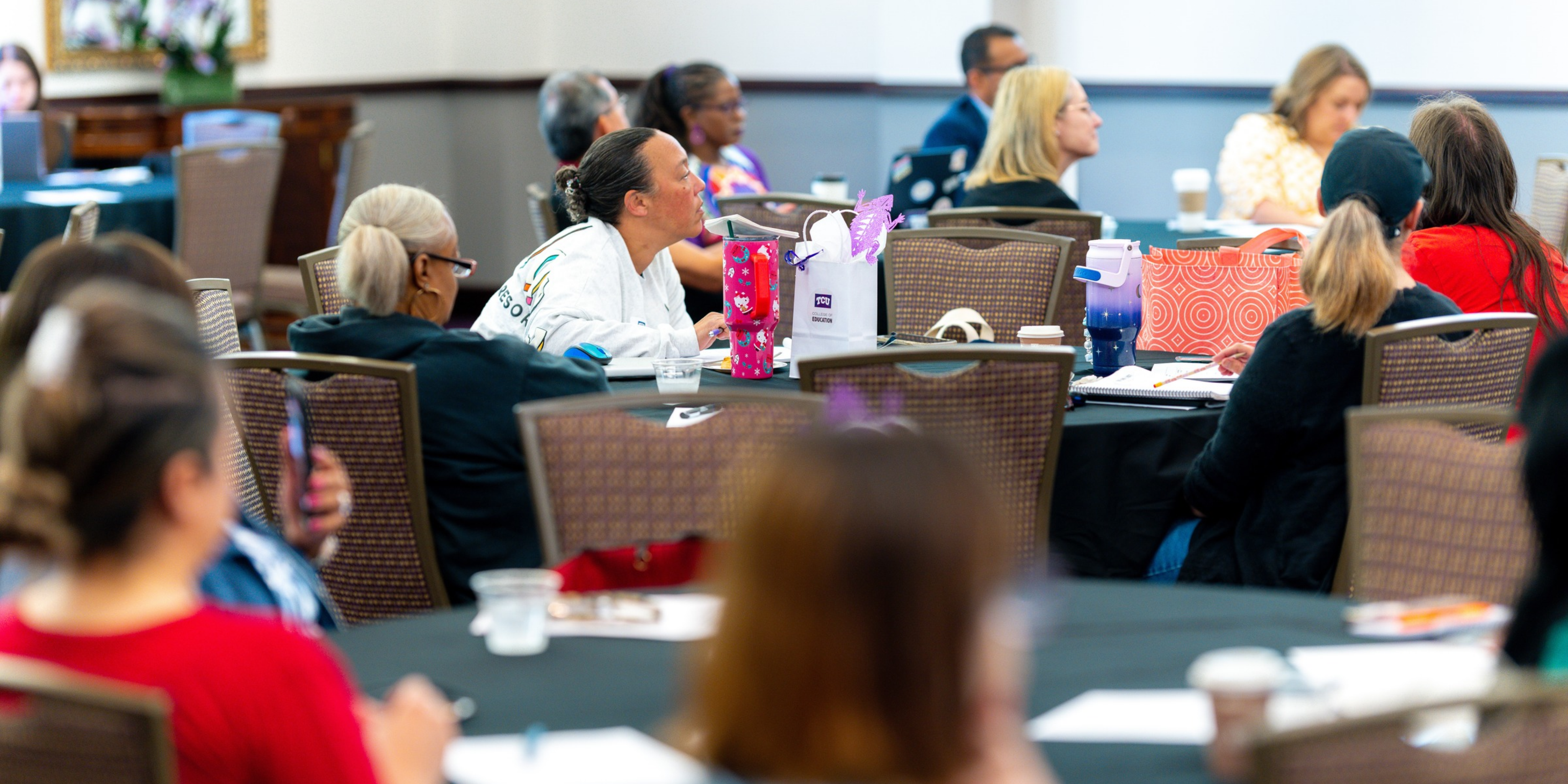A group of people seated at round tables in a conference room, attentively listening to a presentation.
