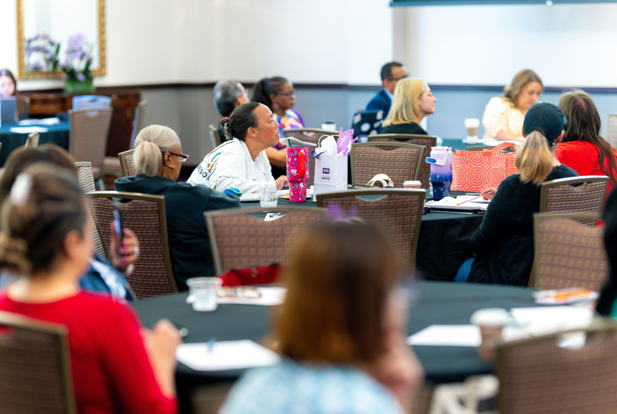A group of people seated at round tables in a conference room, attentively listening to a presentation.