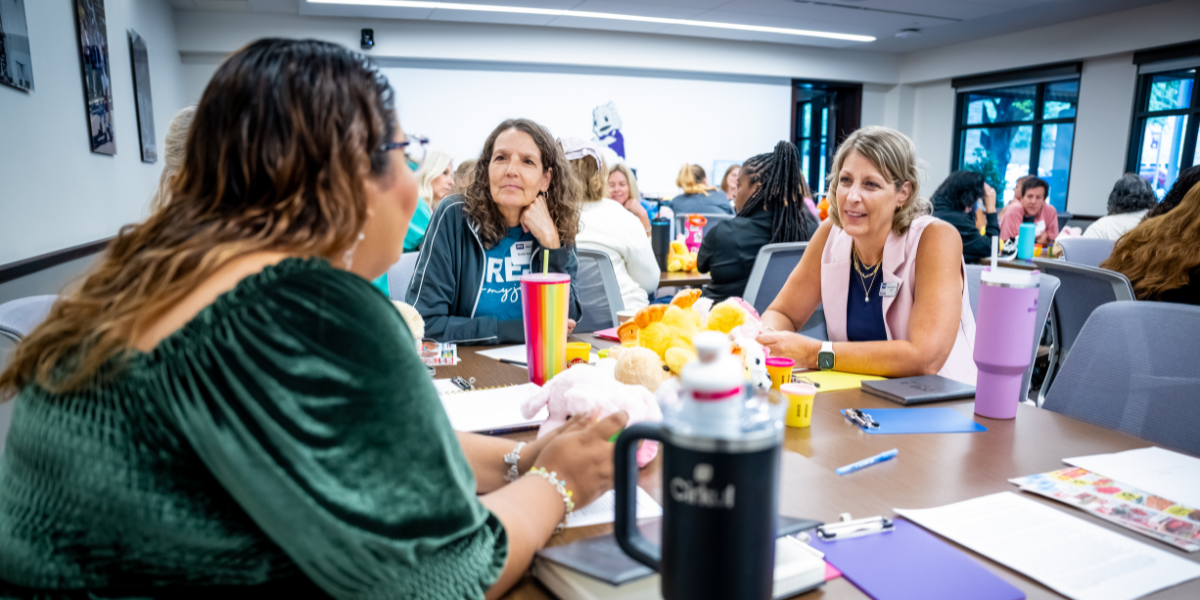 TCU Professor Robin Griffith engaging in conversation with two institute attendees in a meeting room. The table is cluttered with notebooks, colorful mugs, and a variety of stuffed toys.