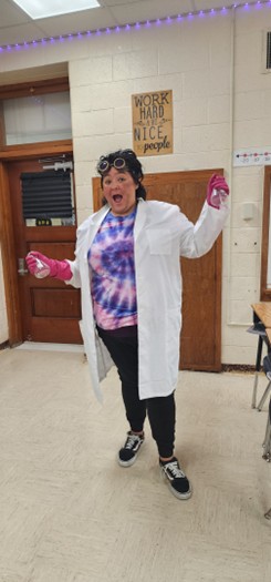 Naomi Gutierrez in a white lab coat and wearing steampunk goggles on her head. Her face is made up to looked singed for her "Mad Scientist" Halloween costume.