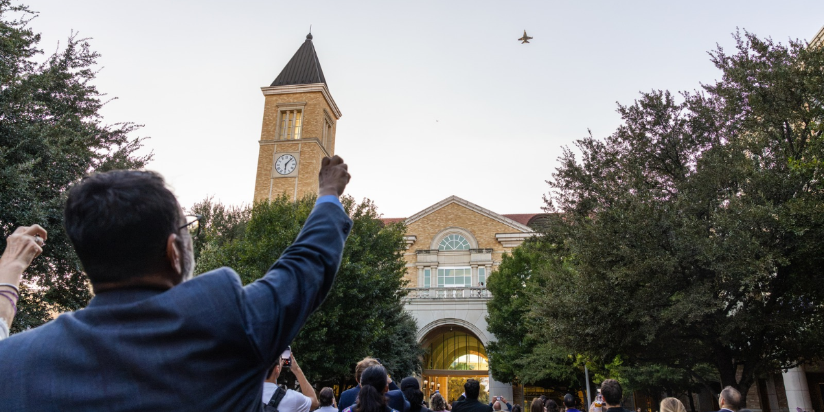 Crowd gathered outside Texas Christian University building with a clock tower, watching a jet fly overhead.