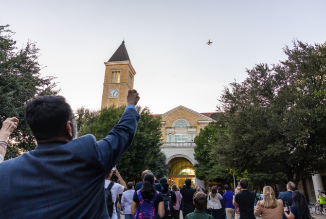 Crowd gathered outside Texas Christian University building with a clock tower, watching a jet fly overhead.