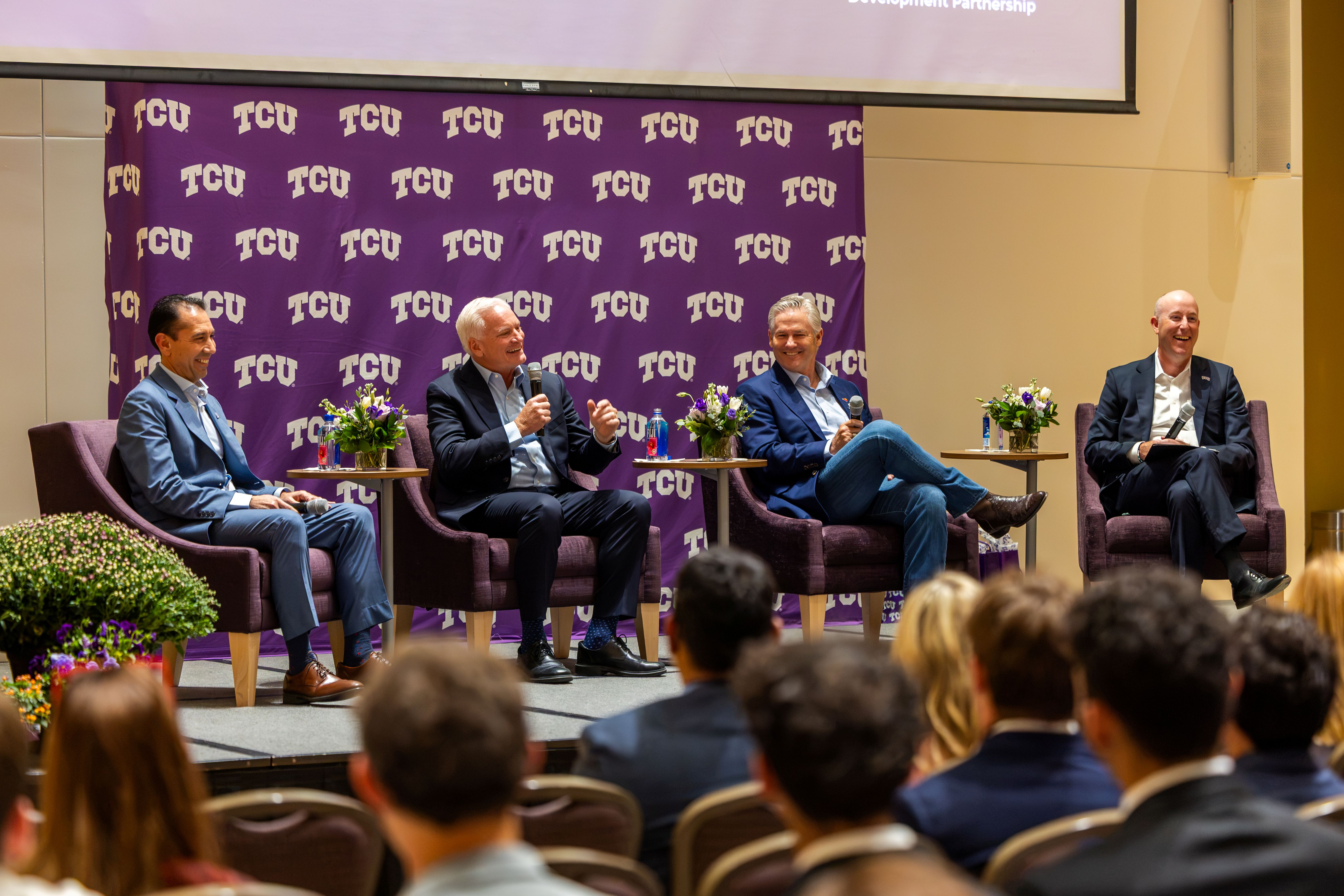 A panel of four individuals is seated on stage with microphones, engaged in discussion. The background features a TCU-branded banner. An audience is visible in the foreground.