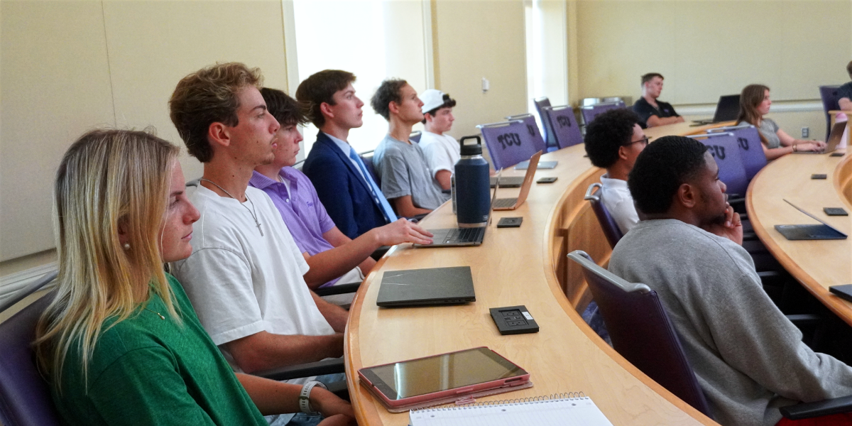 A classroom with attentive students seated in a curved row, facing forward. Some have laptops, notebooks, or tablets on the desk. "TCU" is visible on the seating.