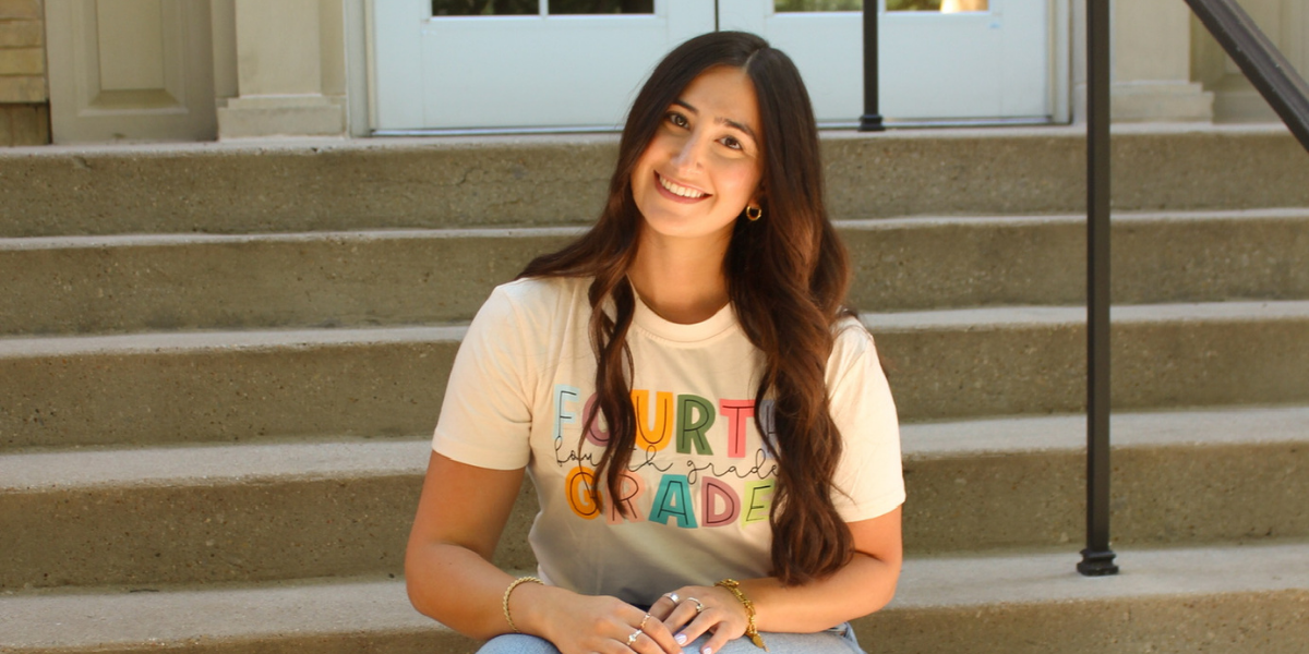 A person sits on outdoor steps in front of a building with glass-paneled doors. They are wearing a "Fourth Grade" t-shirt, jeans, and white shoes, smiling while sitting with hands clasped on their lap.