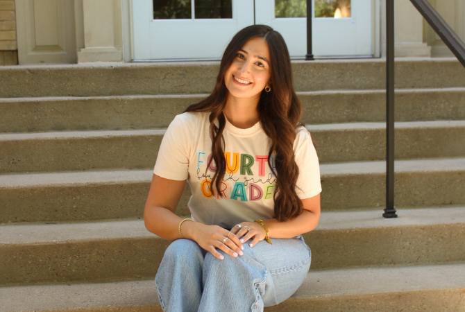 A person sits on outdoor steps in front of a building with glass-paneled doors. They are wearing a 