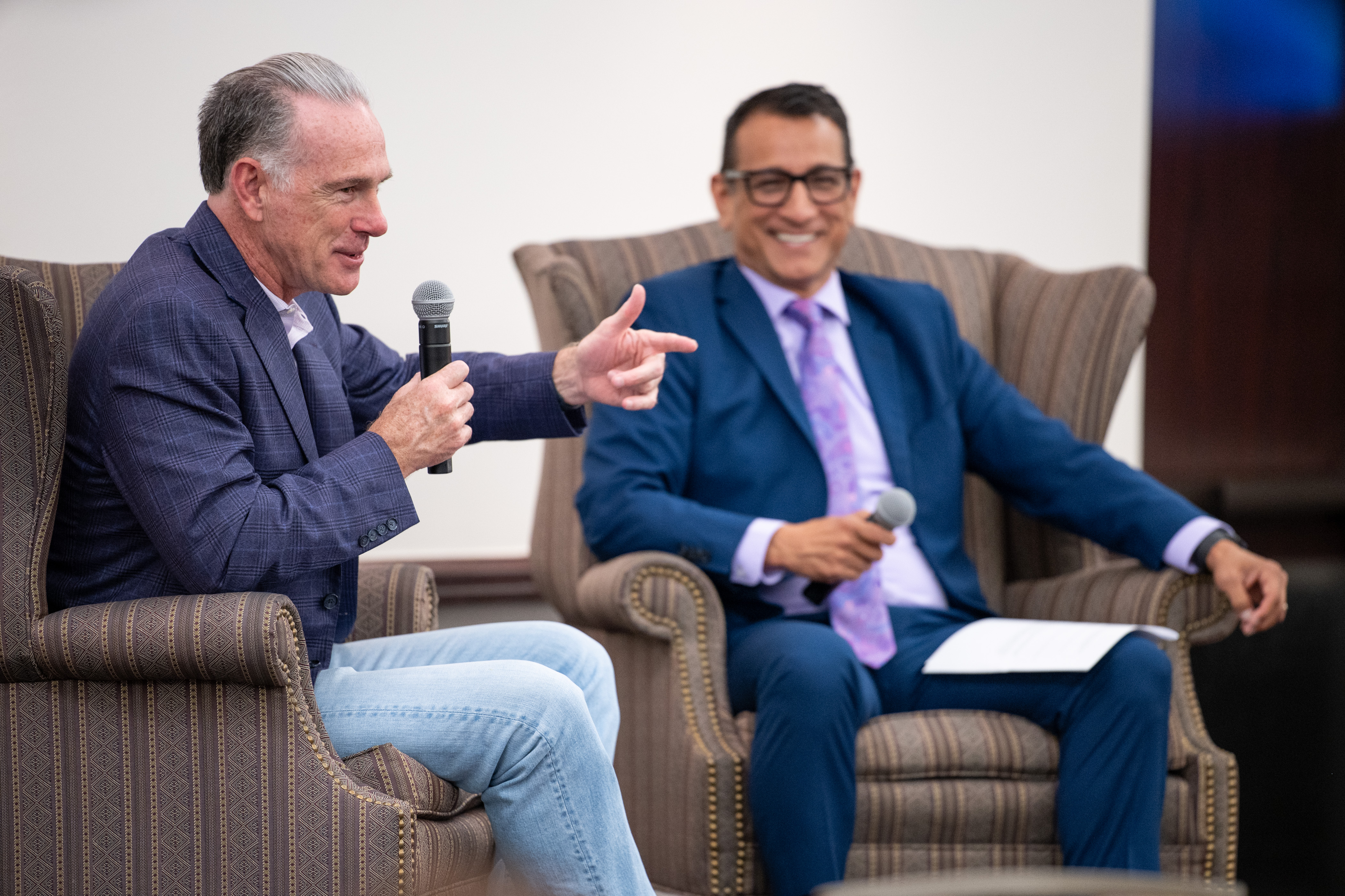 TCU Men's Basketball Coach sitting with TCU College of Education Dean Frank Hernandez. Both sit on wingback chairs and wear suits. Coach Dixon holds a microphone and points toward the audience off-screen.