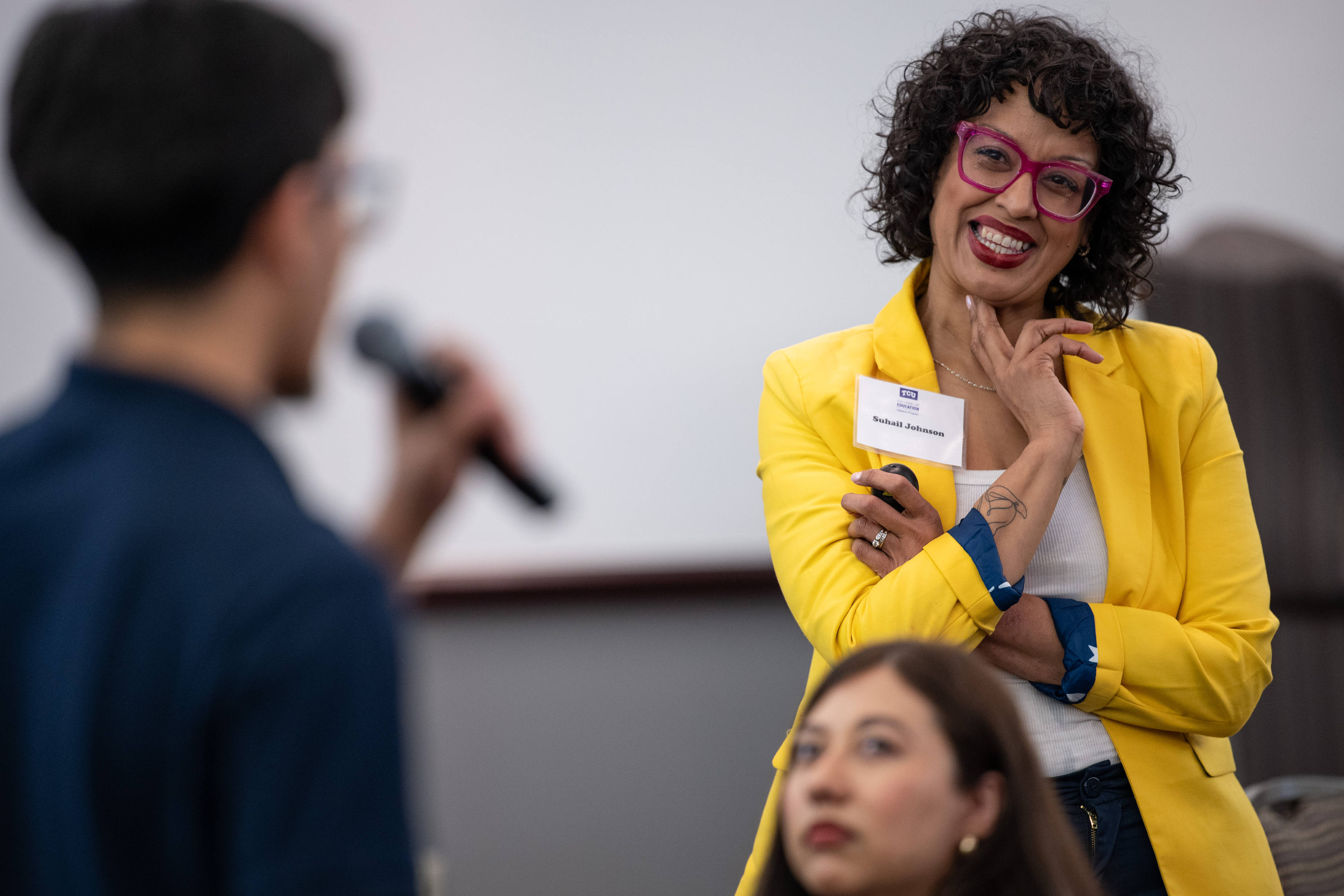 Suhail Johnson, wearing a yellow blazer and a nametag, looks on as a MAESTRO Conference attendee speaks during her interactive presentation. The speaker is blurred in the foreground.