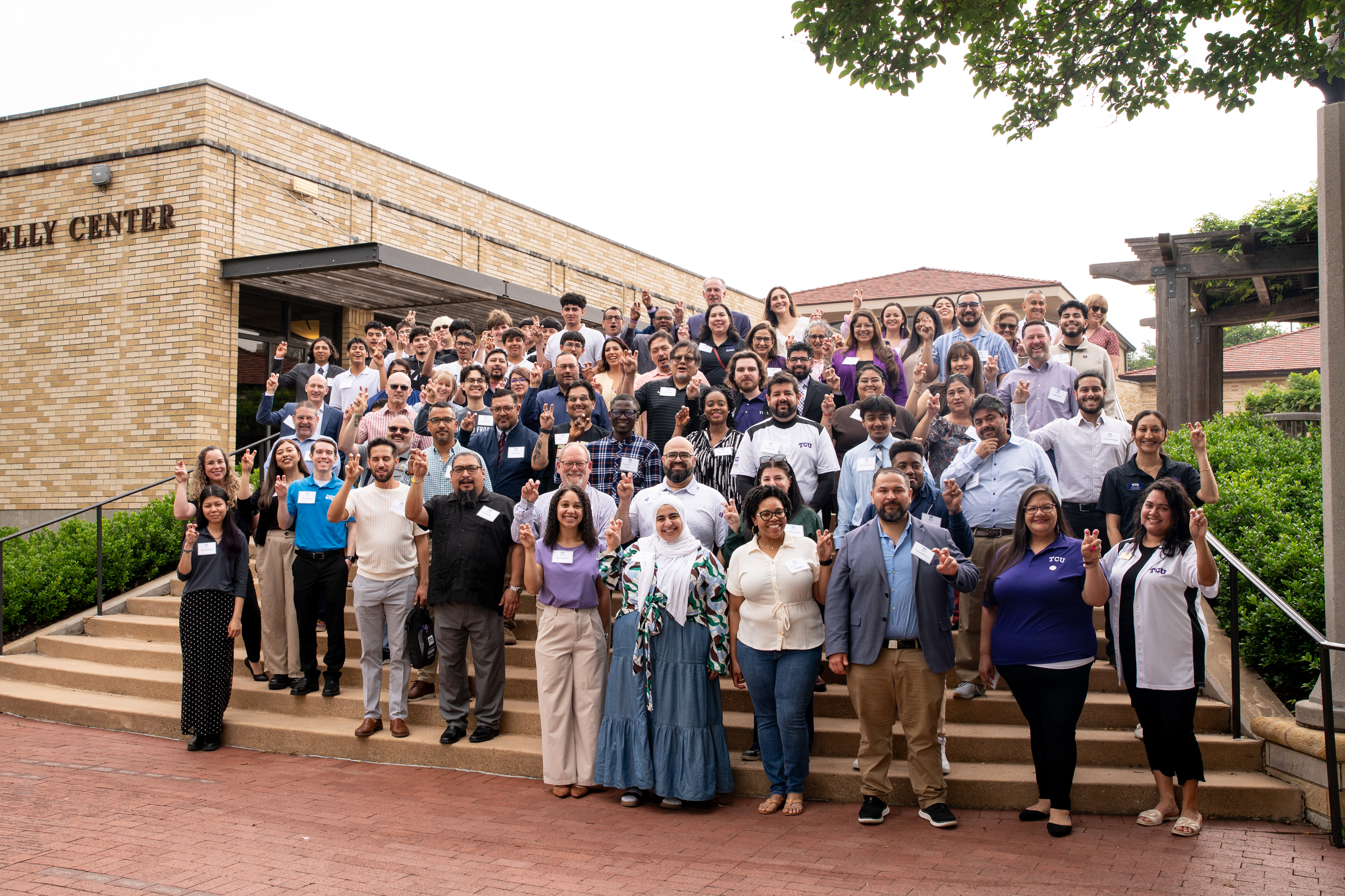 A large, diverse group of people standing on steps outside the Dee J. Kelley Alumni Center. They are posing for a group photo, wearing name tags and business casual clothing. Trees, greenery and a pergola are visible in the background.