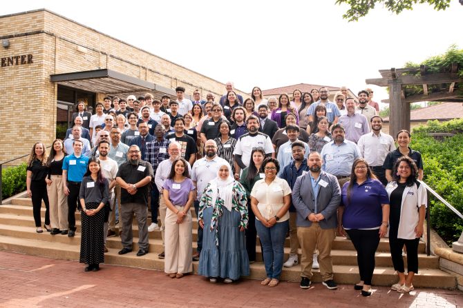 A large, diverse group of people standing on steps outside the Dee J. Kelley Alumni Center. They are posing for a group photo, wearing name tags and business casual clothing. Trees, greenery and a pergola are visible in the background.