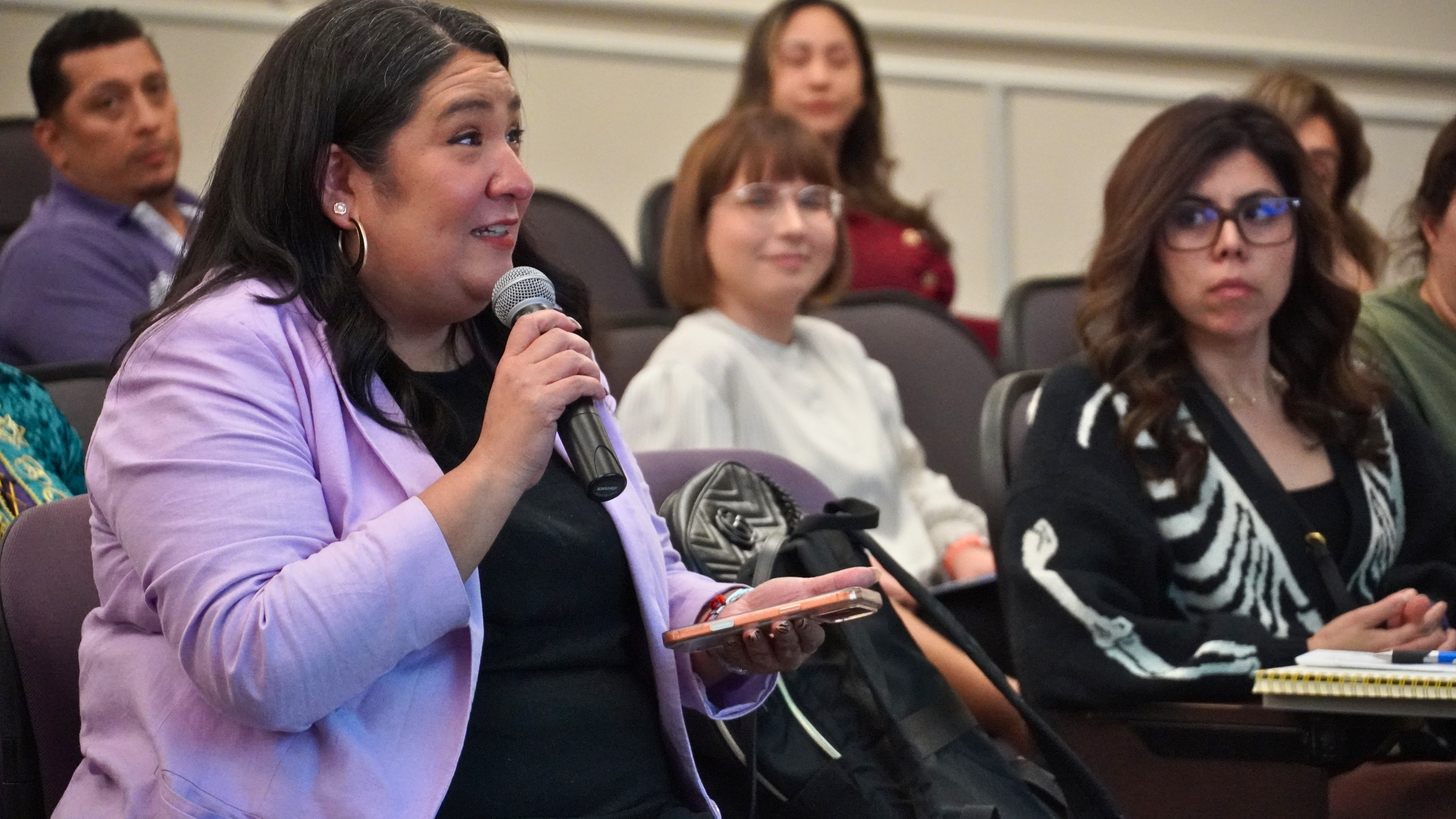 A person in a purple blazer is speaking into a microphone in a seated audience. Other attendees are listening attentively, holding notebooks and looking towards the speaker.