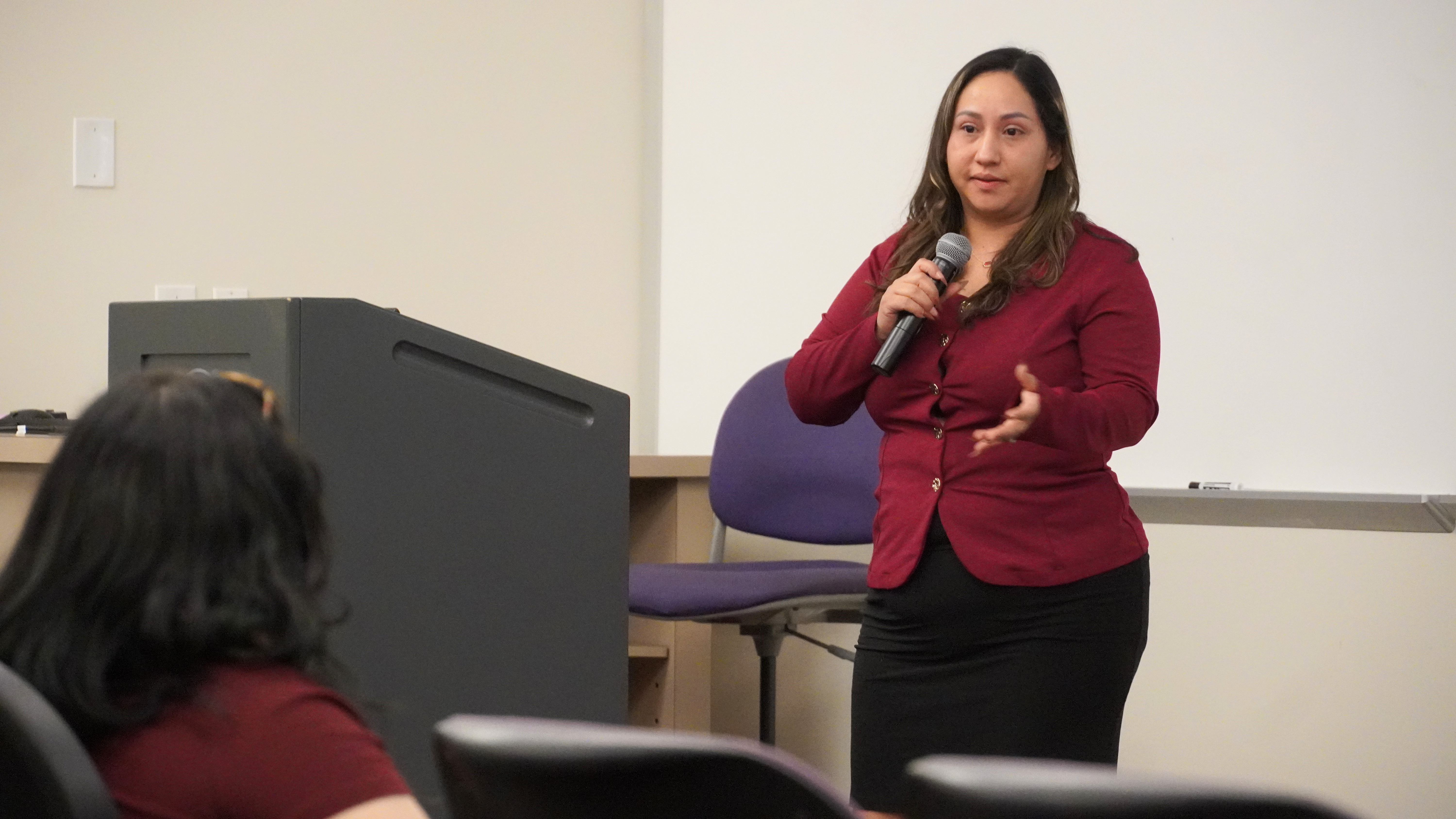 A person speaking into a microphone stands at the front of a room, addressing an audience. A large computer station is to the left. The setting appears to be a classroom or seminar.
