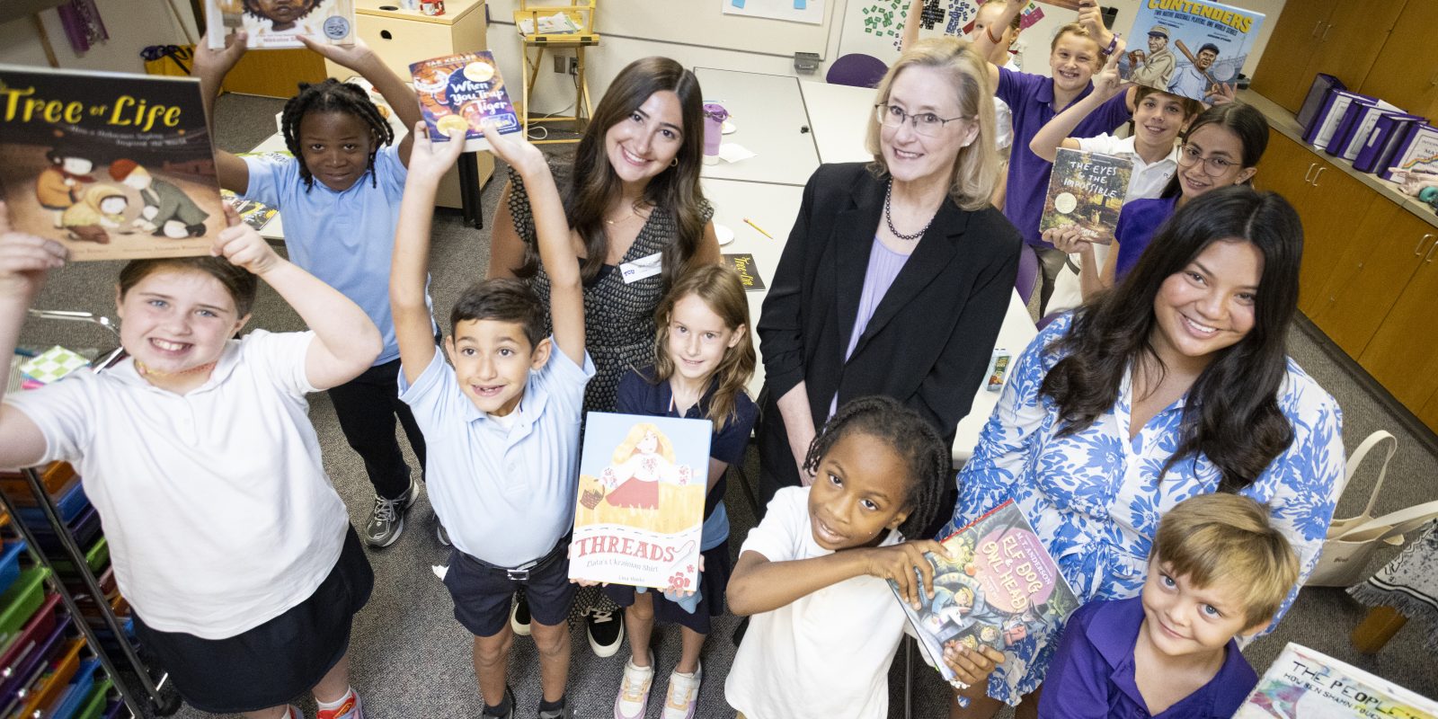 A group of people, including children and two adults, are smiling and holding books in a classroom. The books displayed include titles like "Tree of Life" and "Threads." Shelves filled with educational materials are visible in the background.