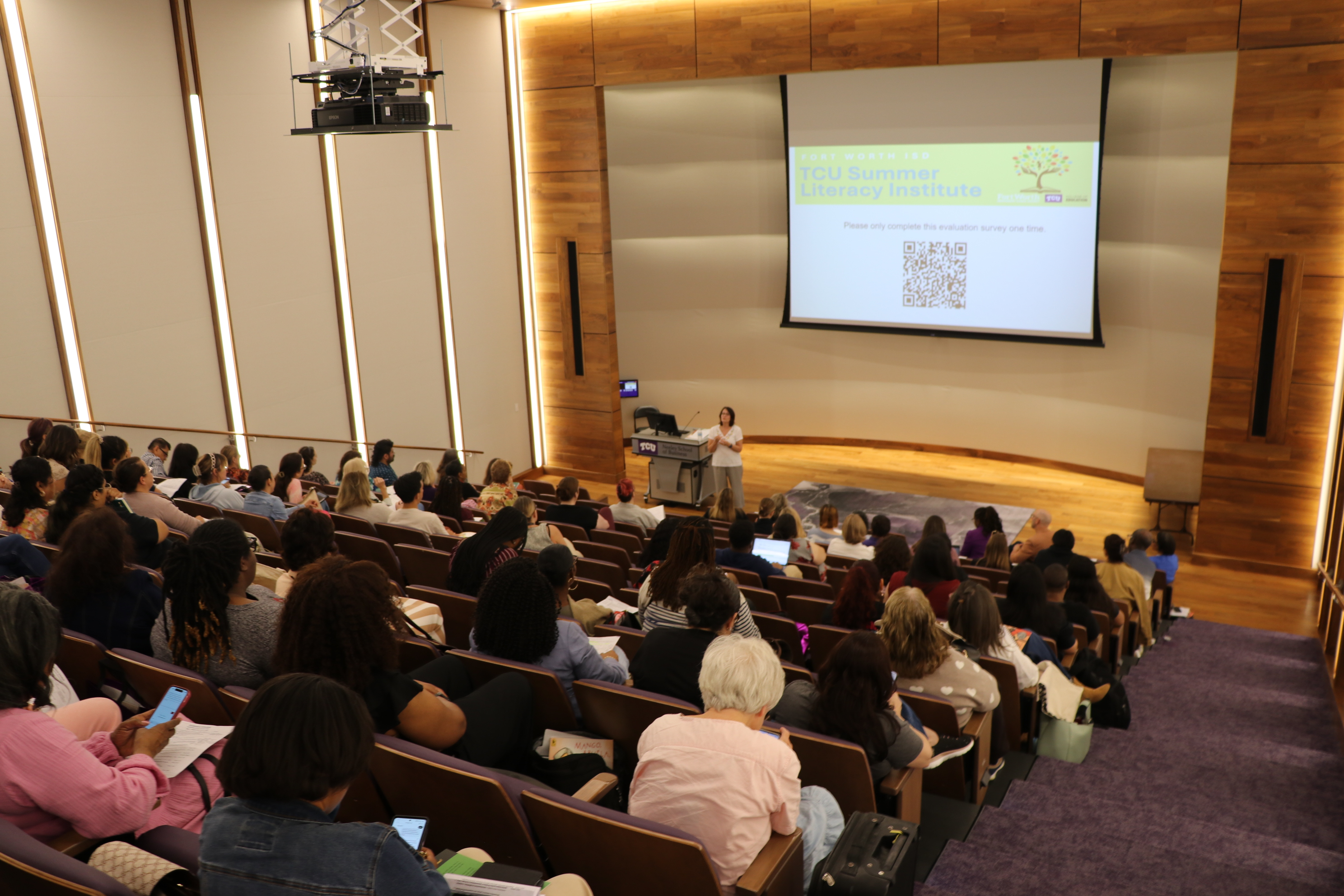 A lecture hall filled with attendees watching a presentation at the "TCU Summer Literacy Institute." The presenter stands at the front beside a screen displaying the event name, a QR code, and a message. The room features wooden paneling and tiered seating.