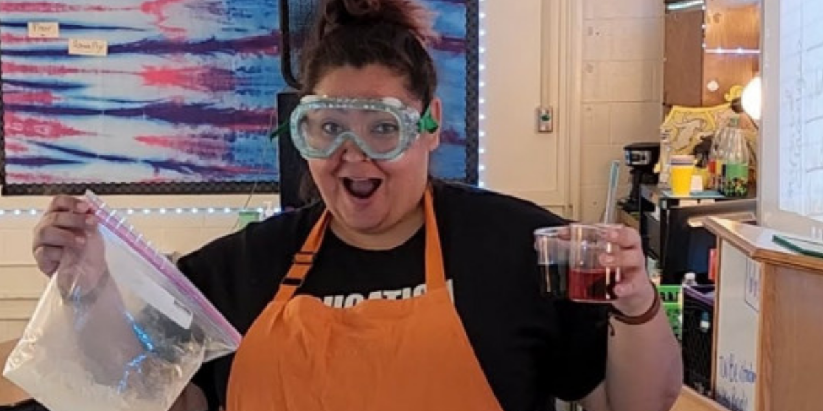 Naomi Gutierrez wearing an orange apron and safety goggles, holding up the components for a science experiment.