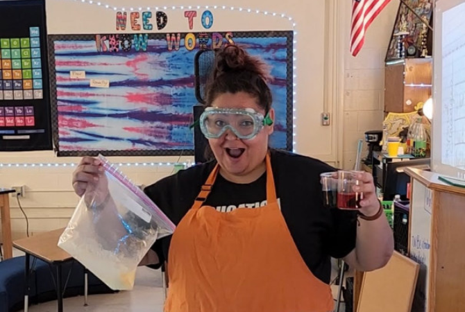 Naomi Gutierrez wearing an orange apron and safety goggles, holding up the components for a science experiment.